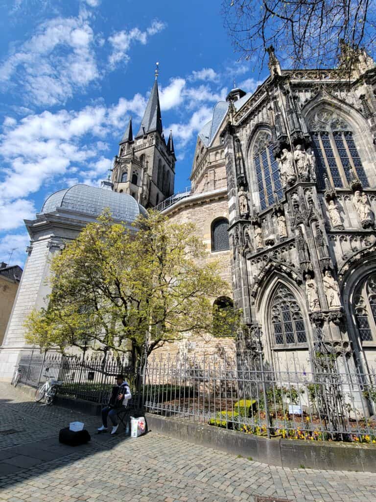 A man sitting on a low wall playing an accordion with an ornate, gothic building with stained glass windows and an intricate spire