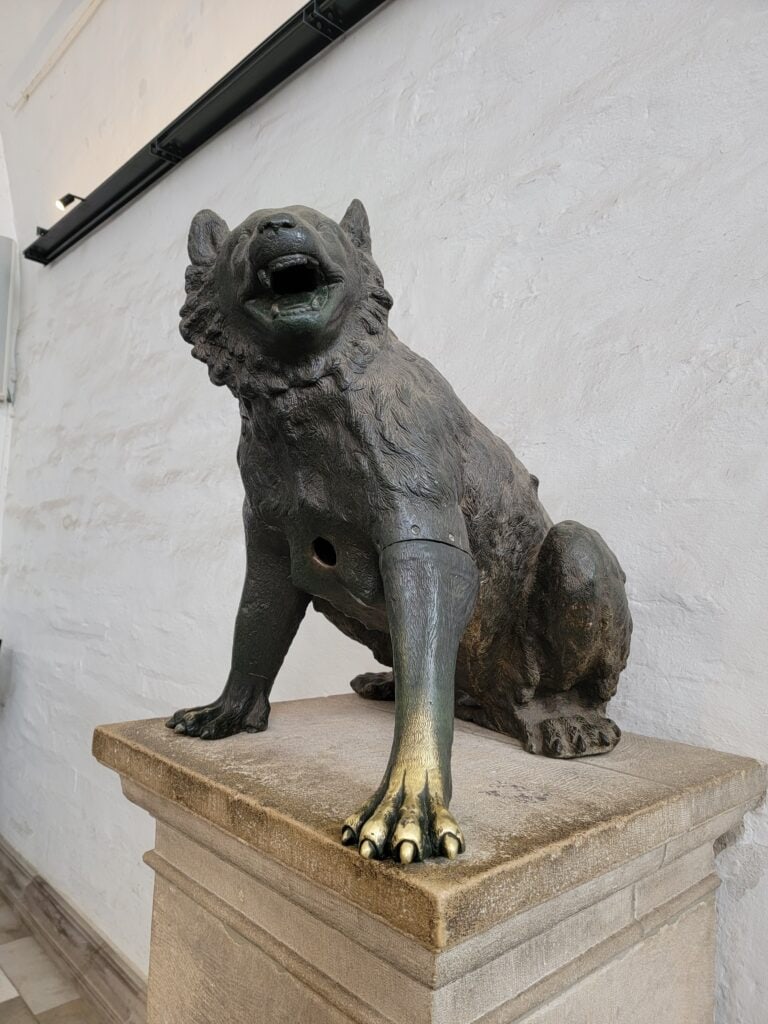 A dark bronze wolf sits, looking up with open mouth. Her front left paw has been rubbed to a shiny yellow brass color