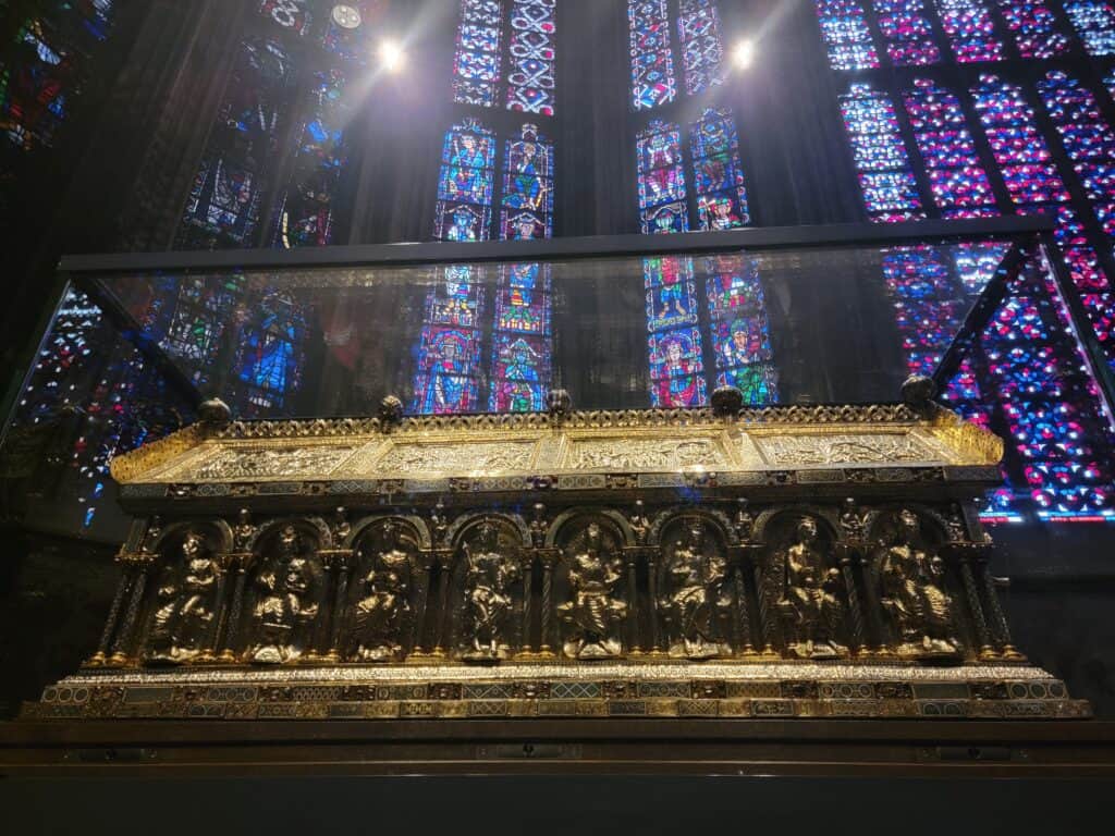 A golden casket with figures on the side, tall columns of blue stained glass behind