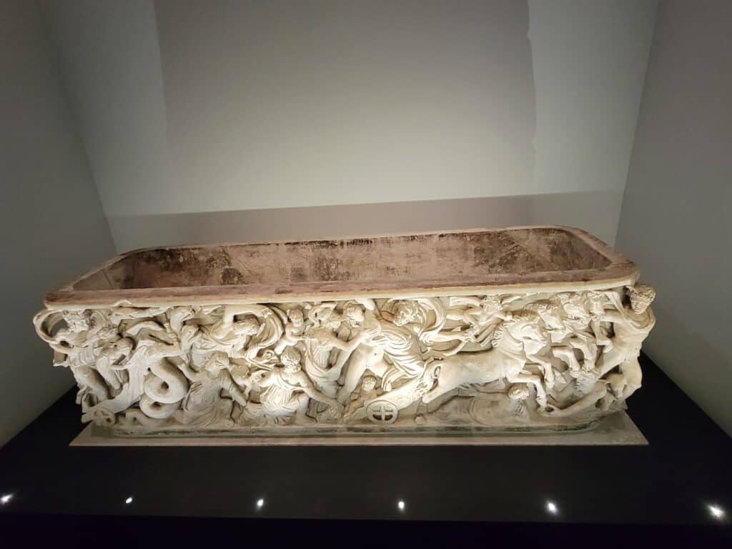 A large white marble sarcophagus with deep carvings of a chariot, horses and people