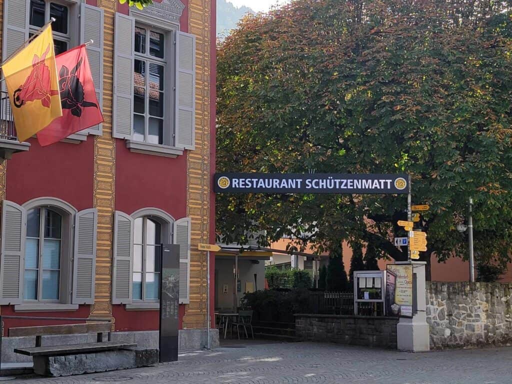 A red building with two flags of cow heads on the left, a banner reading Restaurant Schützenmatt and large trees to the right
