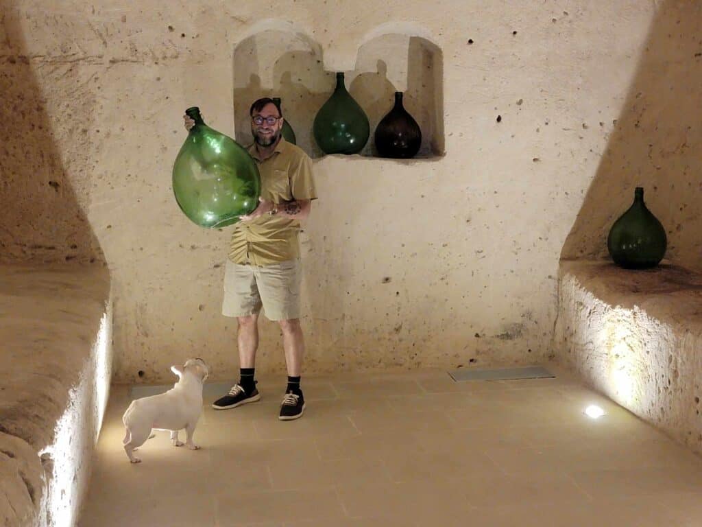 A man in a stone room holds a large green demijohn bottle while a french bulldog looks on.