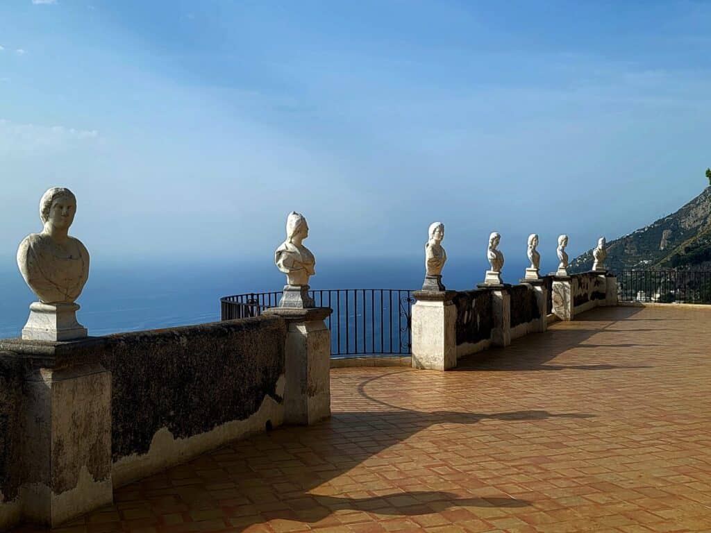 A terazzo terrace lined with white stone busts down its entire length with the blue sea and blue sky behind. 