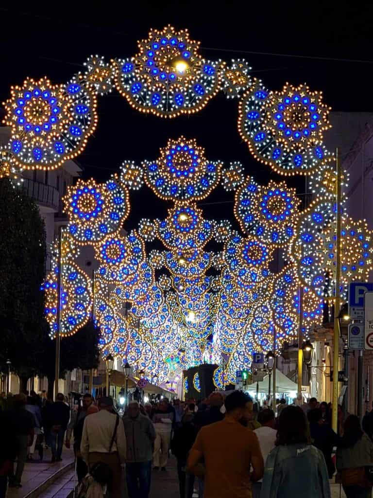 Festive archways of blue and gold flower-shaped lighting panels over a dark street filled with people