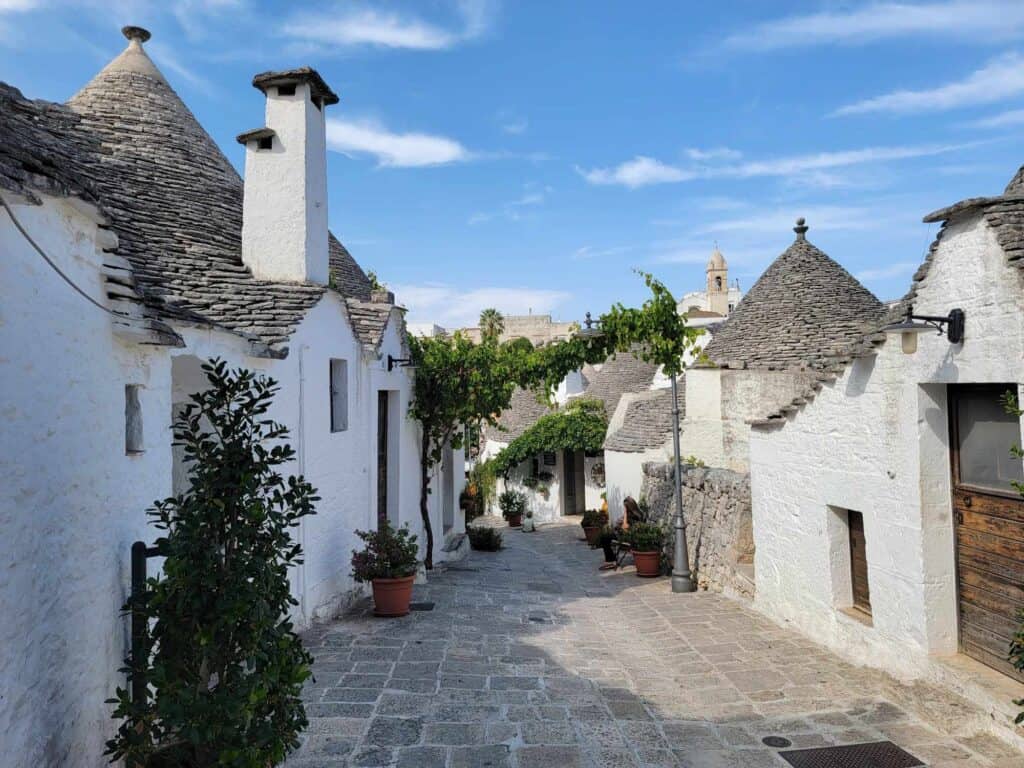 A dark stone road with white trulli huts with conical grey stacked-stone roofs. 