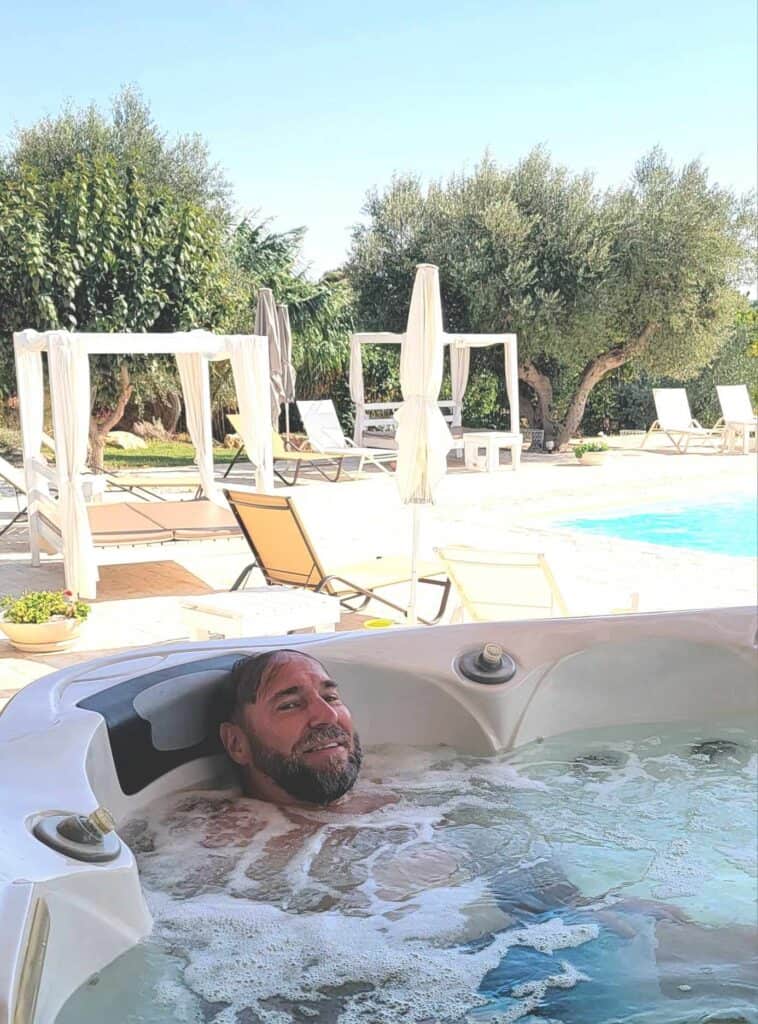 A man almost fully submerged in a hot tub, relaxed and smiling, with lounge chairs and a swimming pool in the background