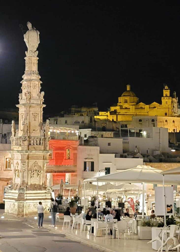 A tall white stone monument and white umbrellas with white tables and chairs filled with diners, a large stone building glows yellow in the background