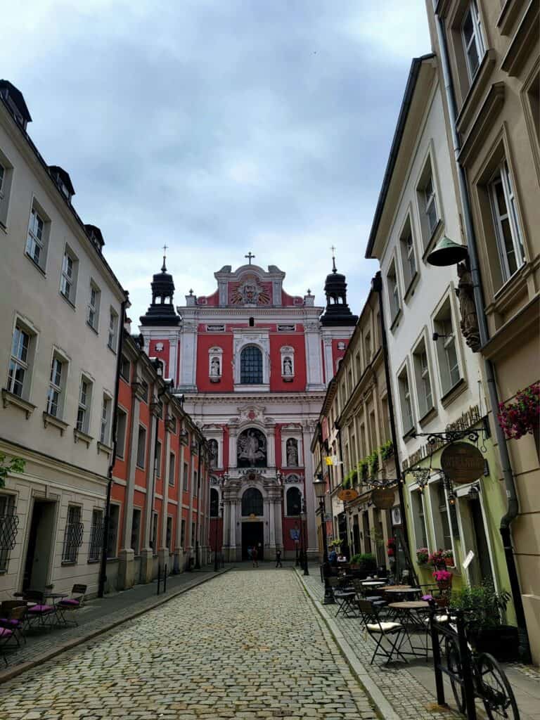 A narrow street with rows of buildings. At the end is the facade of a red and white Baroque church with two small black towers