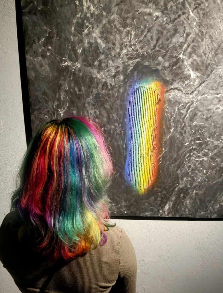 The back of a woman's head with beautiful multicolored hair looking at a painting of a rainbow in the same colors emerging from a dark background