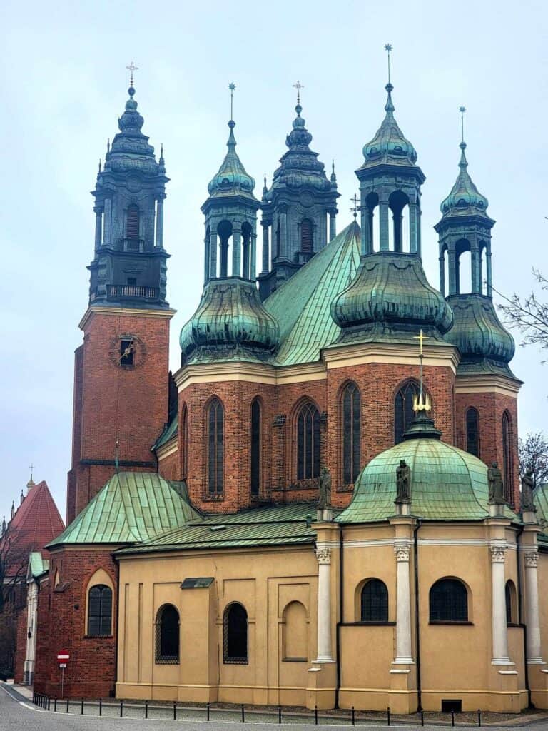 The Poznan cathedral from the outside with five spires and a green copper roof, arched windows, and orange and yellow brick.