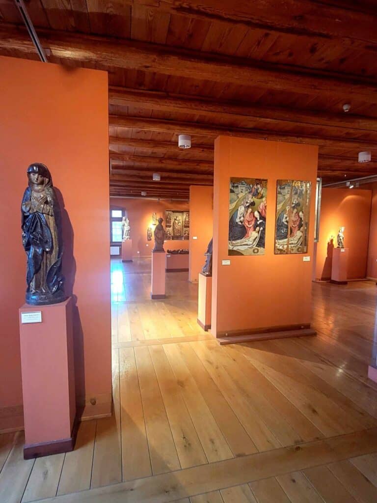 A museum gallery with wooden beamed ceilings and wooden plank floor, with terra cotta colored walls and many wooden Medieval statues and paintings all with religious themes