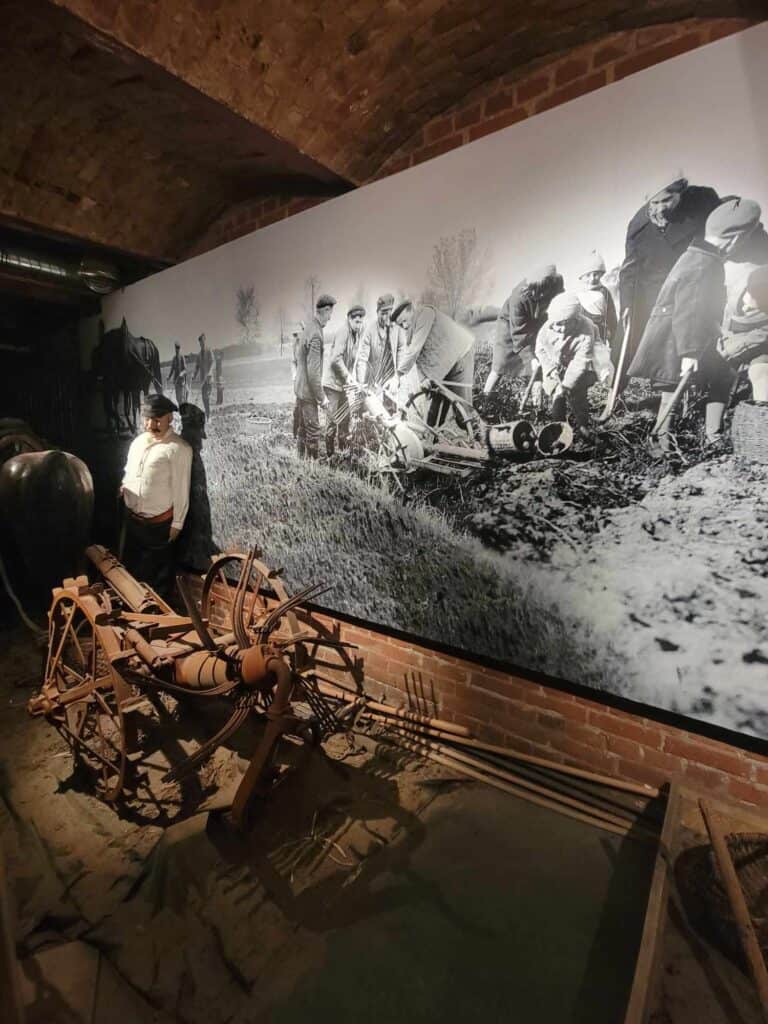 An old wooden farming wagon with wheels in front of a large black and white photo of people using the same wagon