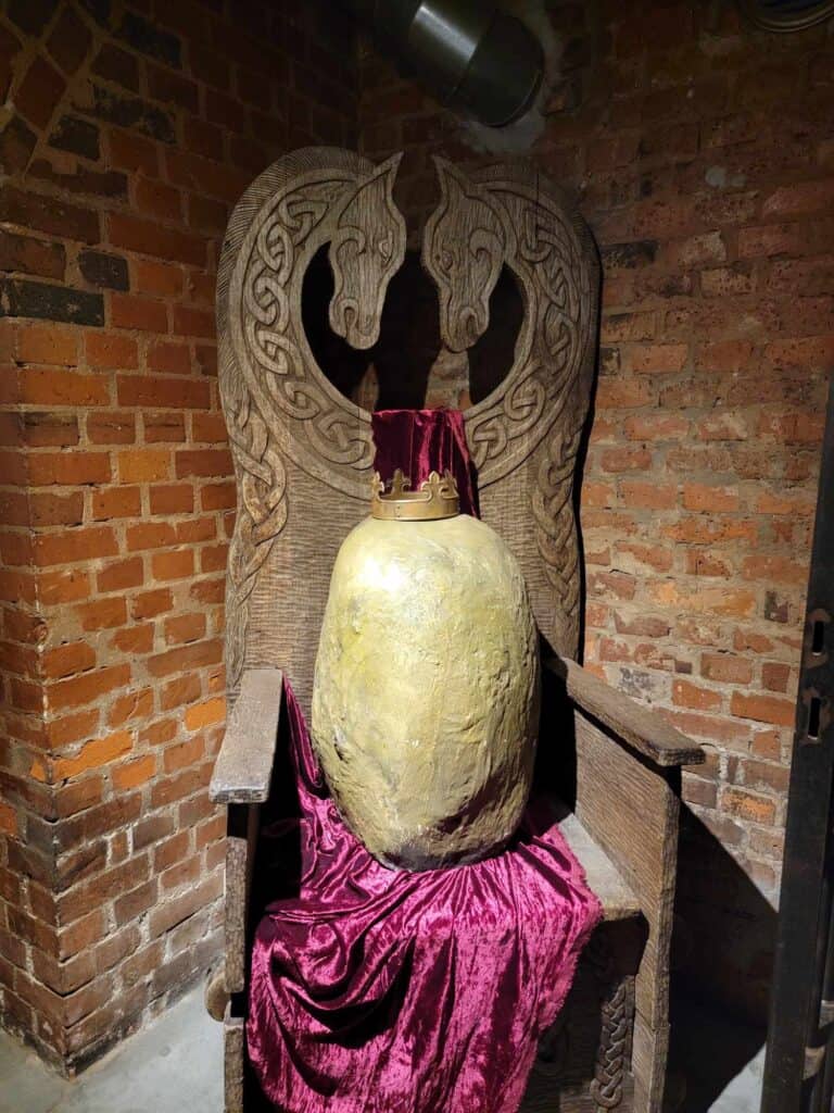 A wooden Celtic looking throne with two horseheads, and an infinity knot border. On it sits an enormous crowned potato sitting on a magenta velvet cloth.