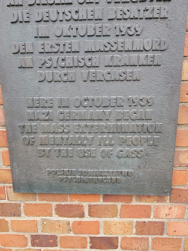 A bronze sign in German and English that reads "Here in October 1939 Nazi Germany began the mass extermination of mentally ill people by the use of gas."