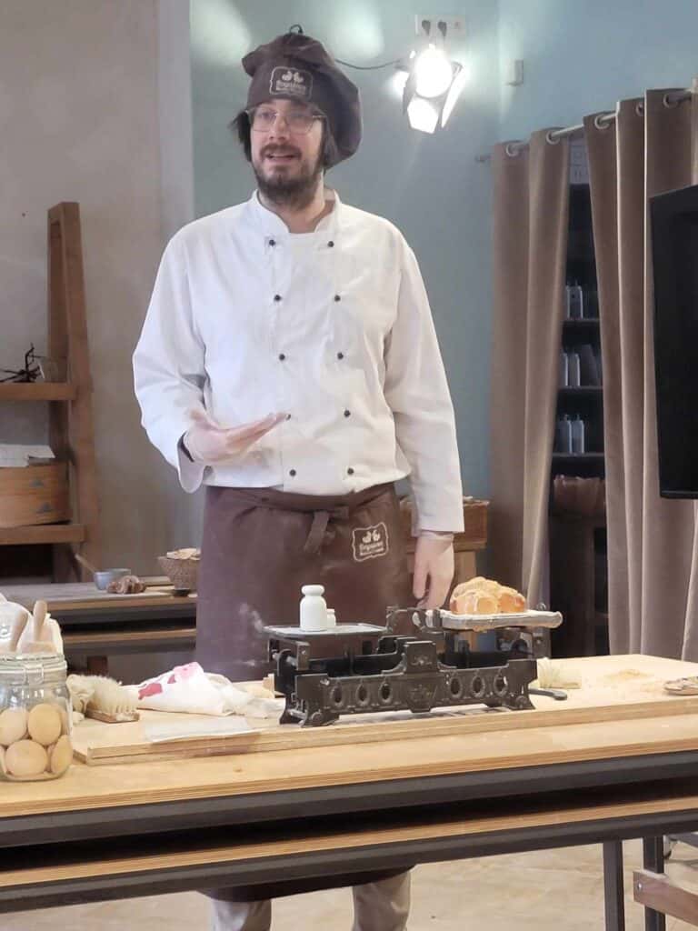 A man in a chef coat and hat weighing a large croissant on a balance scale