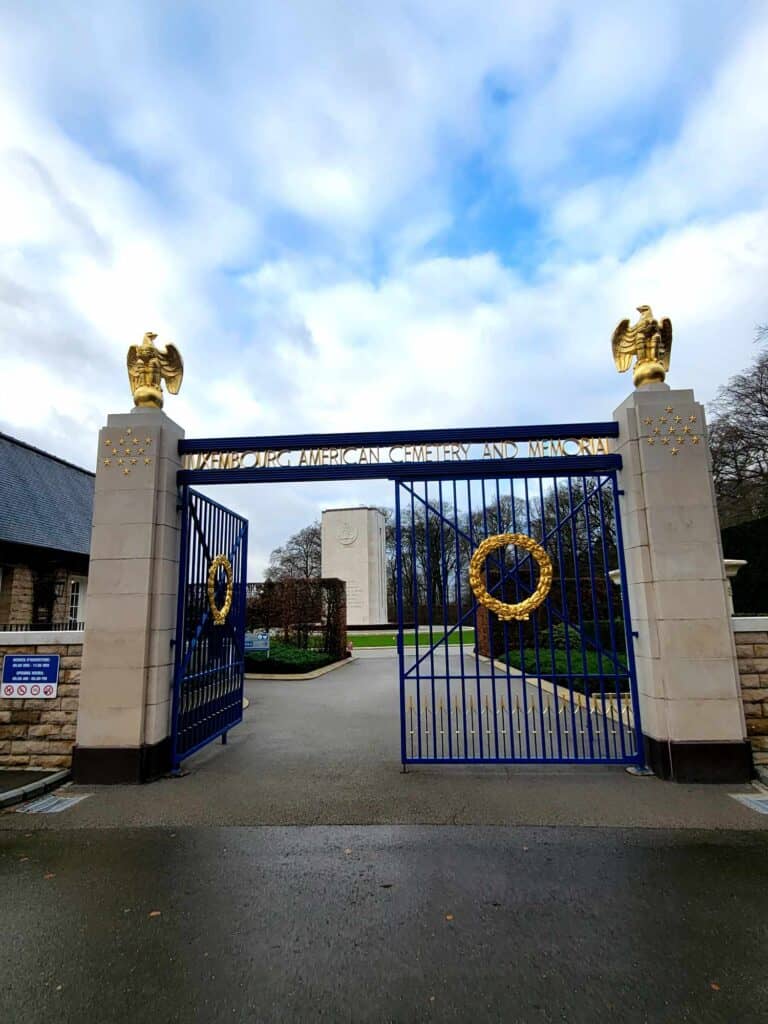 Large stone columns topped with golden eagels support a black gate with two gold wreaths at the entrance of the cemetery where you can visit General Patton's grave