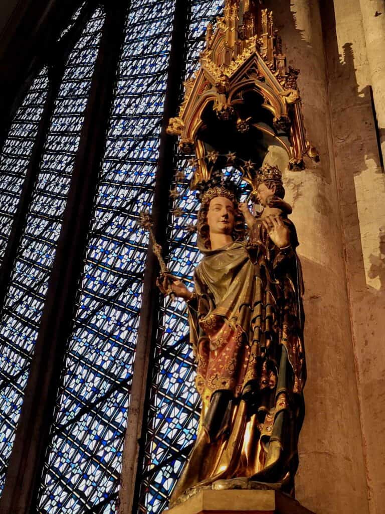 A wooden medieval sculpture of Mary and the baby Jesus next to a column and stained glass window