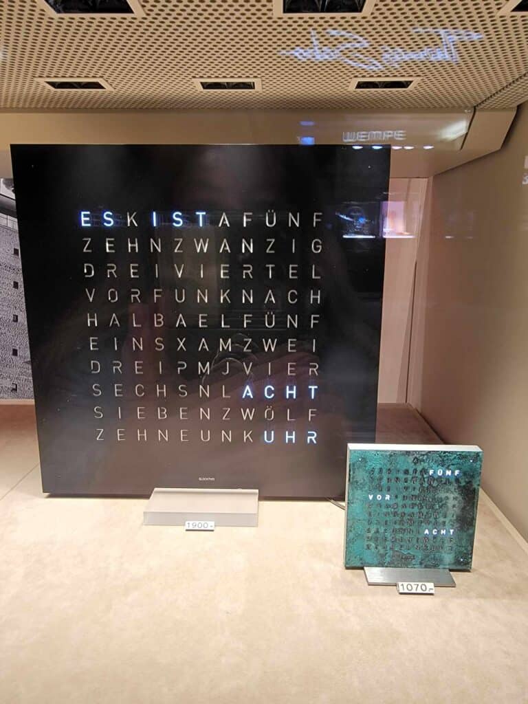 A clock which is square and made up of a panel of cutout letters. The letters are illuminated to reveal the time in words