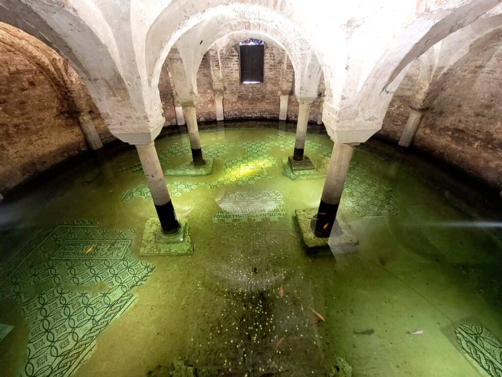 Looking down into a room with white arches held up by support columns and flooded with greenish water revealing fish and partial mosaics on the floor. There are coins in the center and a shuttered window in the back. The water is illuminated from the sides.