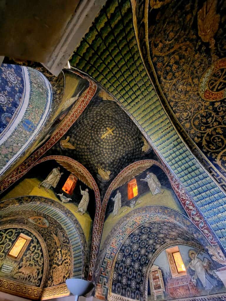 A spectacular vaulted ceiling covered in gold and blue mosaics of people, birds, and stars with a gold cross centered in the dome. If you want to see Ravenna in one day, come here first!