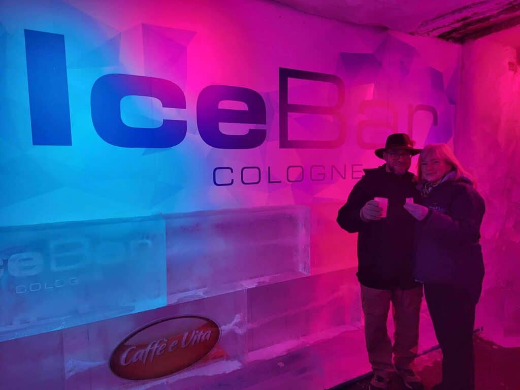 A couple holding drinks in ice glasses in front of a frozen wall illuminated in blue and fuscia that says "Ice Bar Cologne"