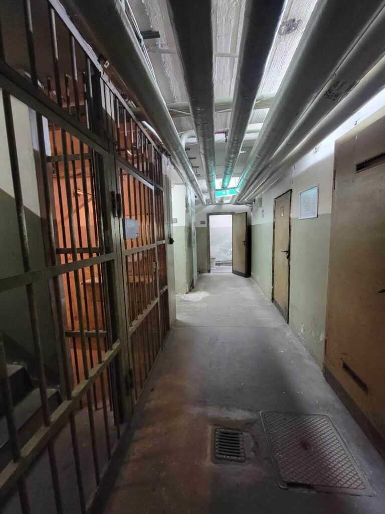 A hallway underground with cells with barred doors on the left side