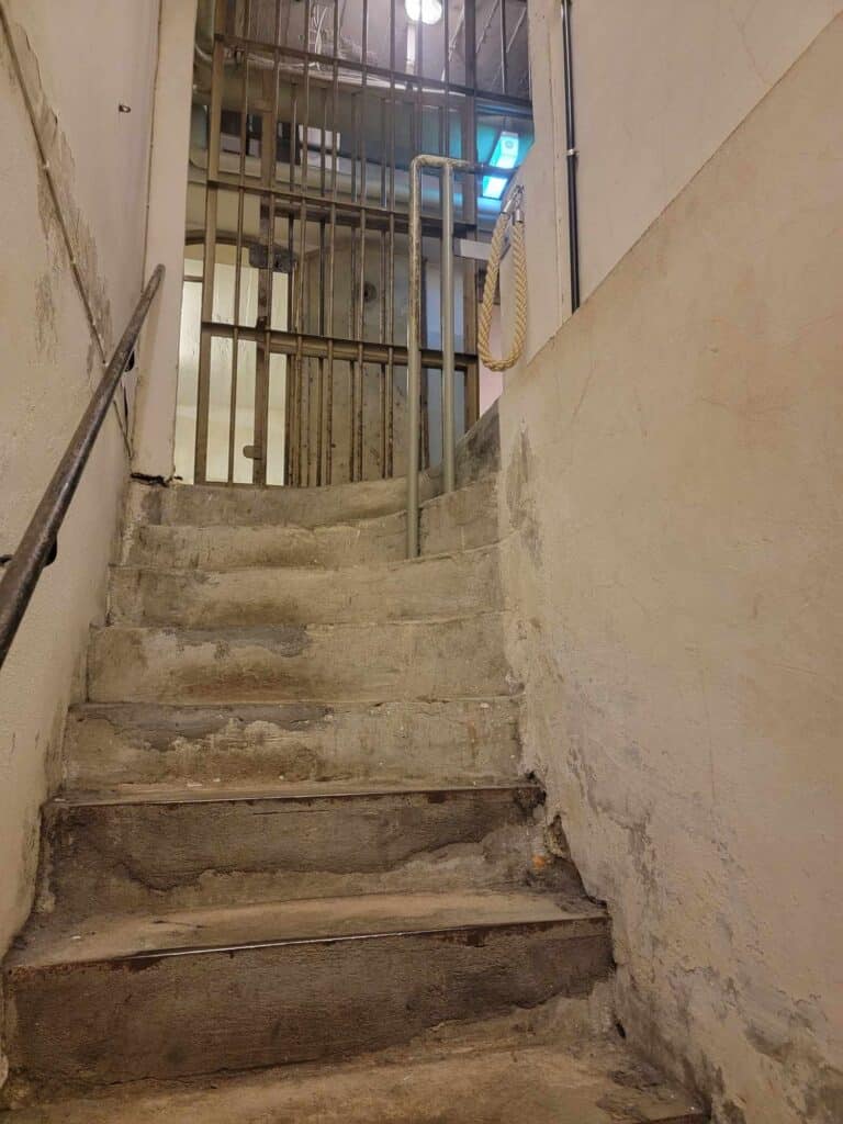 A narrow worn staircase with a barred prison door at the top