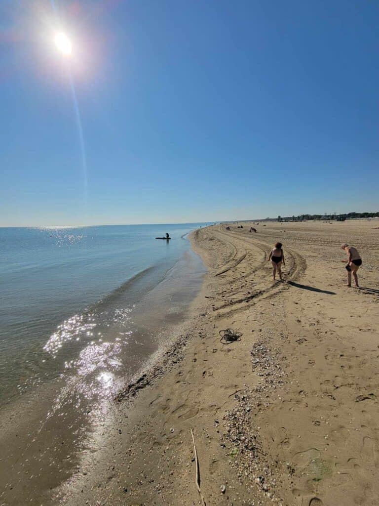 A calm sea with small waves lapping a golden sand shore. Two people are walking on the beach and the sun in the upper left is reflecting on the water and casting shadows