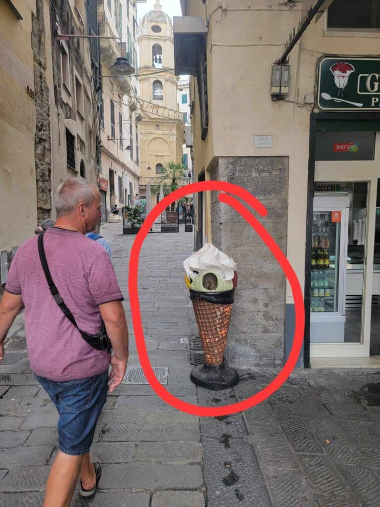 A man walking on a cobblestone street on the left, and on the right a trash can shaped like a vanilla gelato cone with a large hole in the front