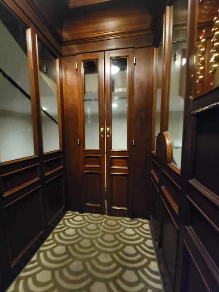 The inside of the elevator all in dark wood with pale green art nouveau style carpet