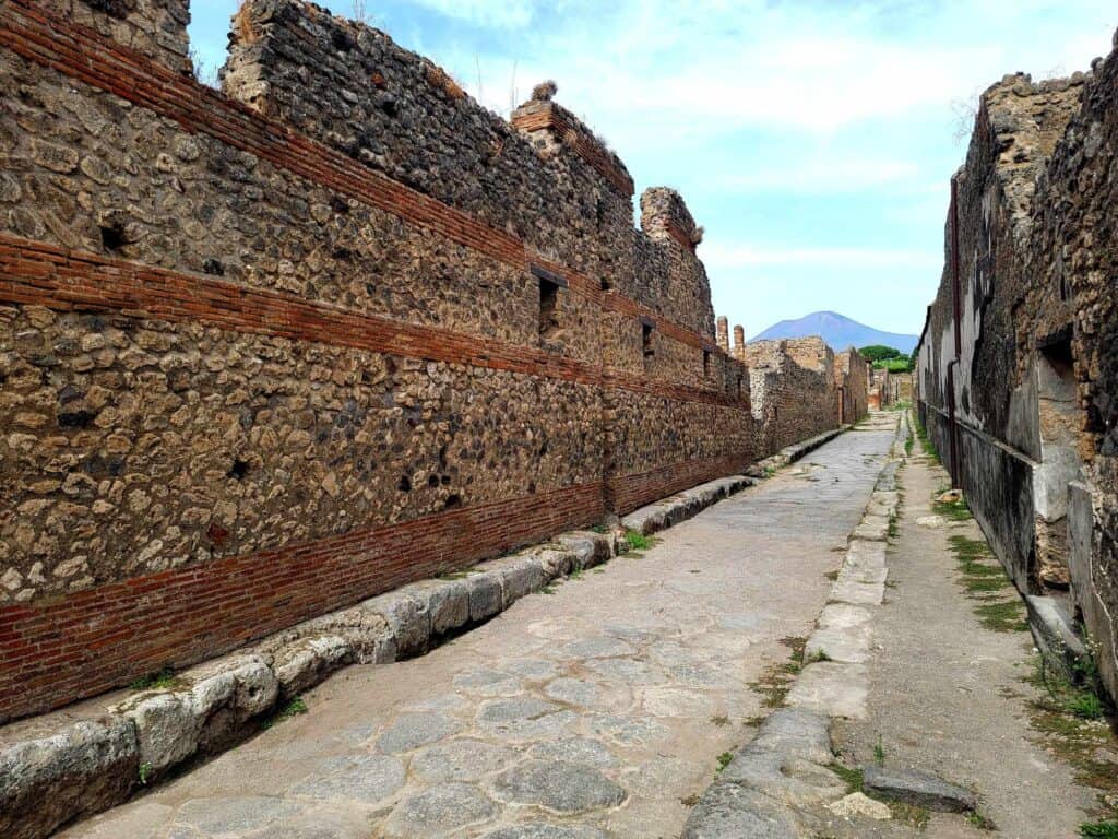 A long road paved with flat rounded stones in Pompeii goes off into the distance with a tall stone wall with stripes of horizontal red brick on the left