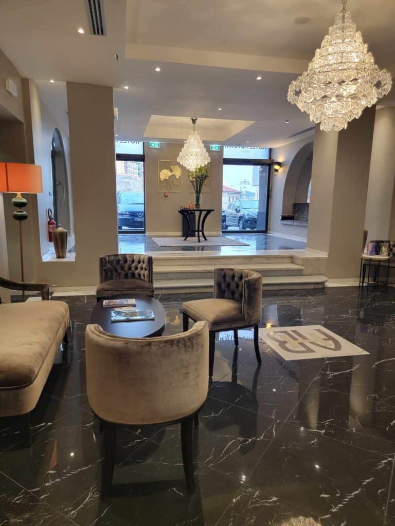 A hotel lobby with marble floors and crystal chandeliers over a sitting area with grey velvet chairs and love seat