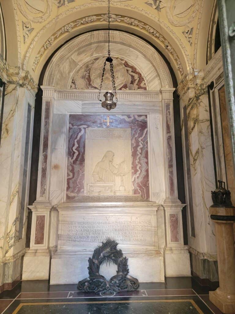 A large marble wall with a relief of Dante holding a book. It houses a large white marble sarcophagus with a carved wreath leaning against it. A lantern hangs down from the ceiling