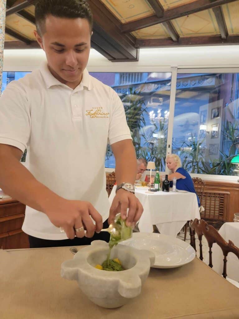 An italian waiter in a white polo shirt pours basil and olive oil into a white marble mortar on a small table