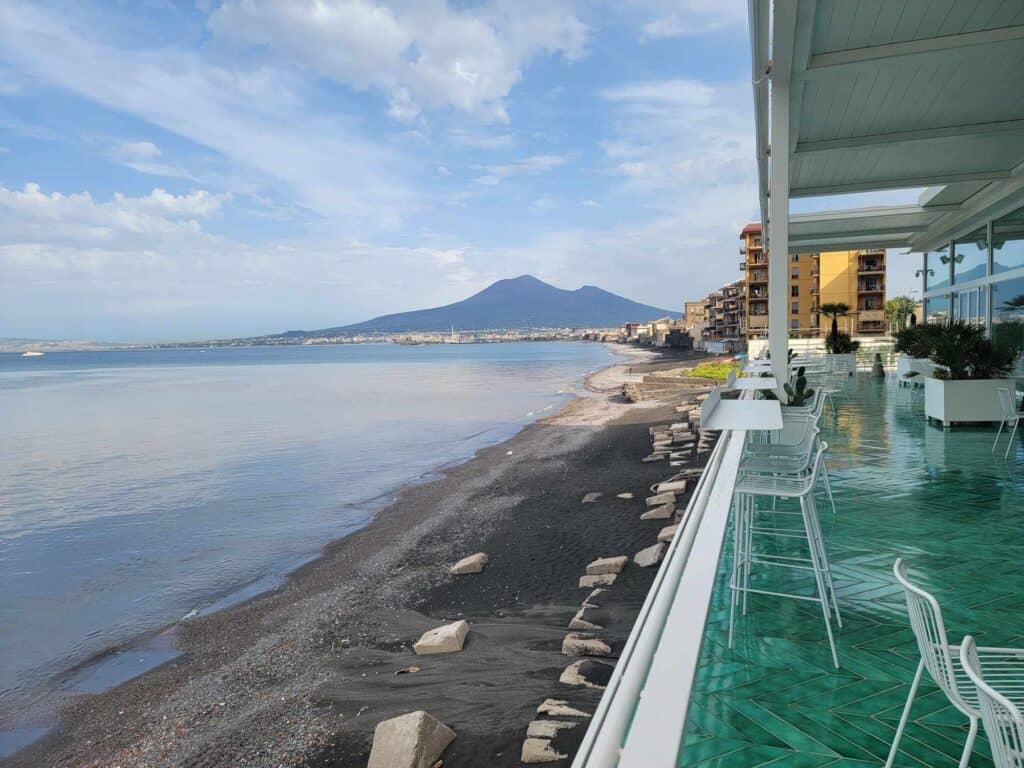 Mount Vesuvius in the background with the very calm Bay of Naples on the left, a ribbon of dark volcanic sand with pale rocks on it, and to the right a bright aquamarine tiled balcony