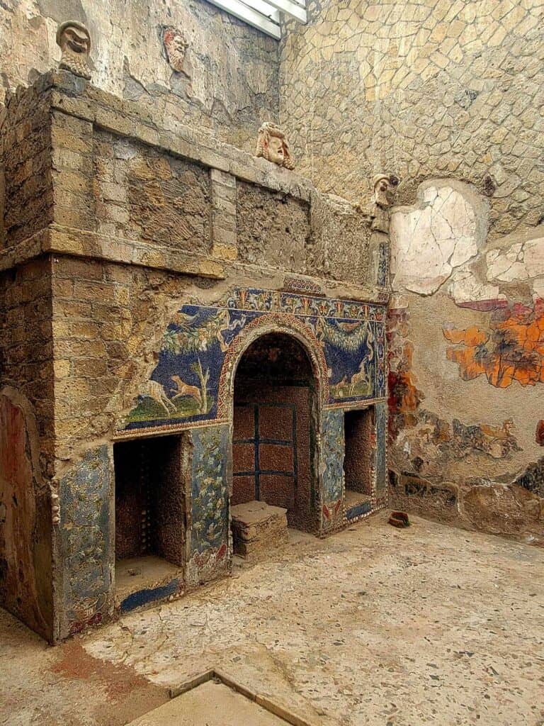 Inside a stone building is a massive fireplace with stone masks above in the Roman style. The front of the fireplace has a partial mosaic - blue with trees and animals