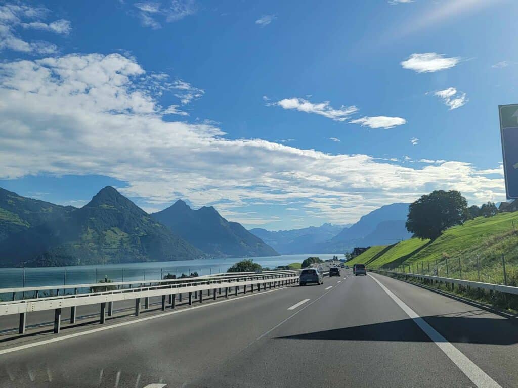 A highway with cars and a long blue lake and mountains on the left.