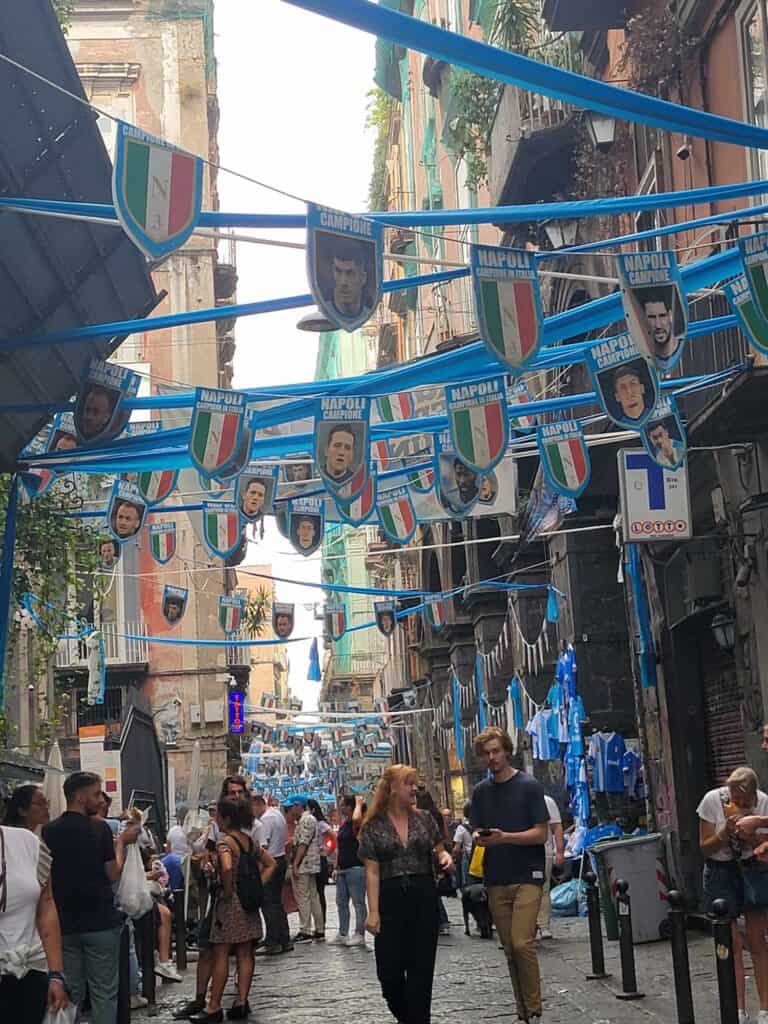 A dark and crowded street draped with blue banners displaying Neopolitan soccer players and Italian flags