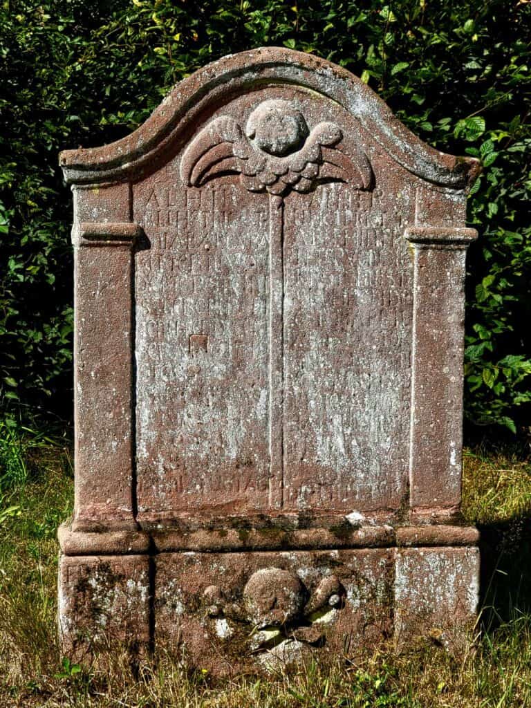 A reddish tombstone with the writing obscured by lichen - an angel at the top and a skull and crossed bones at the base