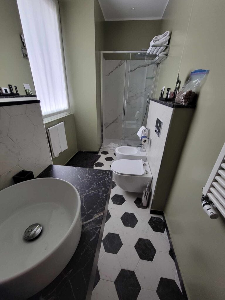 A long thin bathroom with a white basin sink, white fixtures, and hexagonal black and white tiles. A marble shower at the back