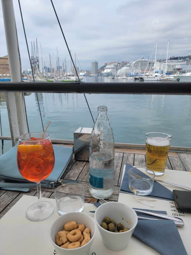 A rustic wooden table on a deck overlooking a harbor is set with an Aperol Spritz, a glass of beer, two glasses of sparkling water, a bowl of taralli snacks, and another of olives