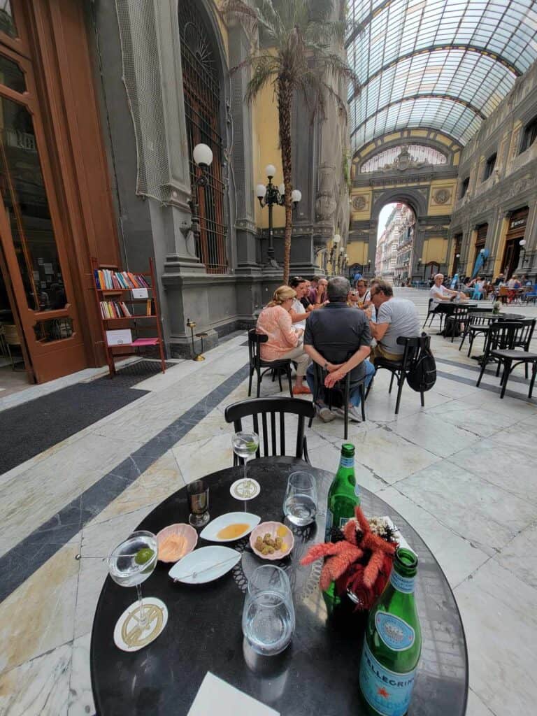 A table with two martinis, two sparkling waters, and a bowl of olives in a covered piazza with a white marble floor and another table with four people sitting in the background