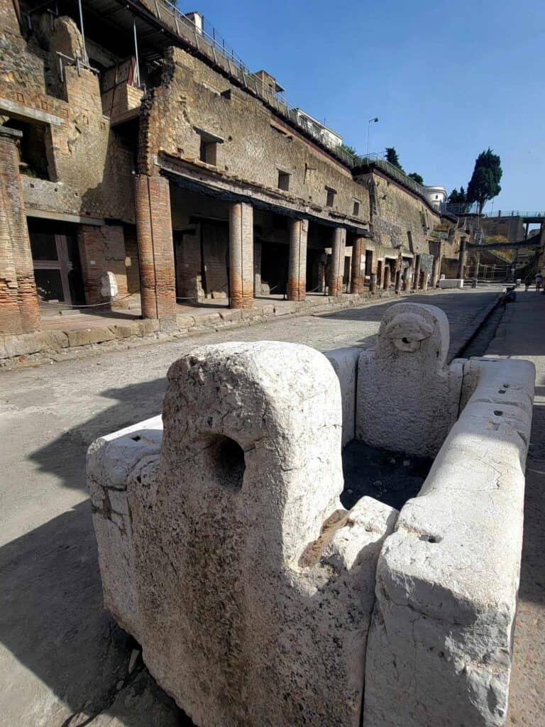 An ancient storefront in Herculaneum with a promenade supported by columns and small windows above. A white stone trough in the foreground which used to be a well