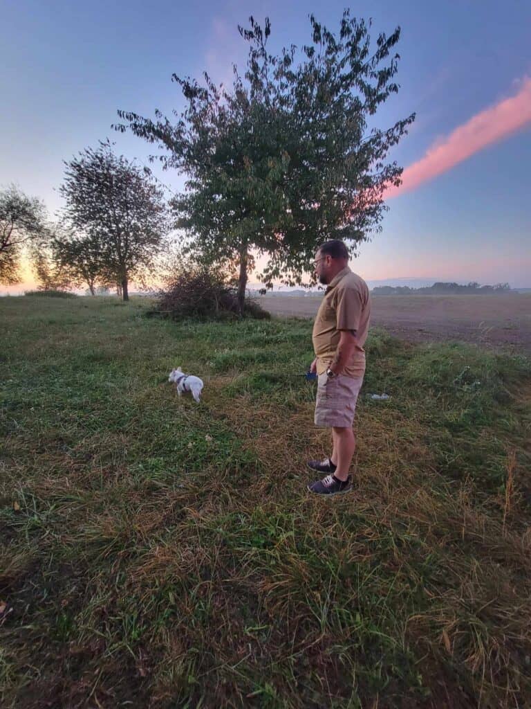 A man and a white french bulldog standing in a large rural field as the sun comes up