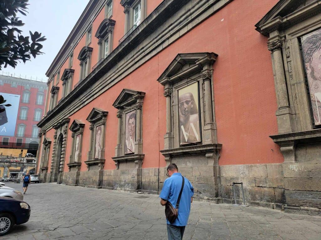 A man looking down, walking past a reddish two story building with large images of Roman paintings and sculptures