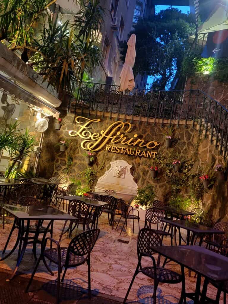 Eight black metal tables surrounded by chairs on a terazzo patio at night with moody uplighting