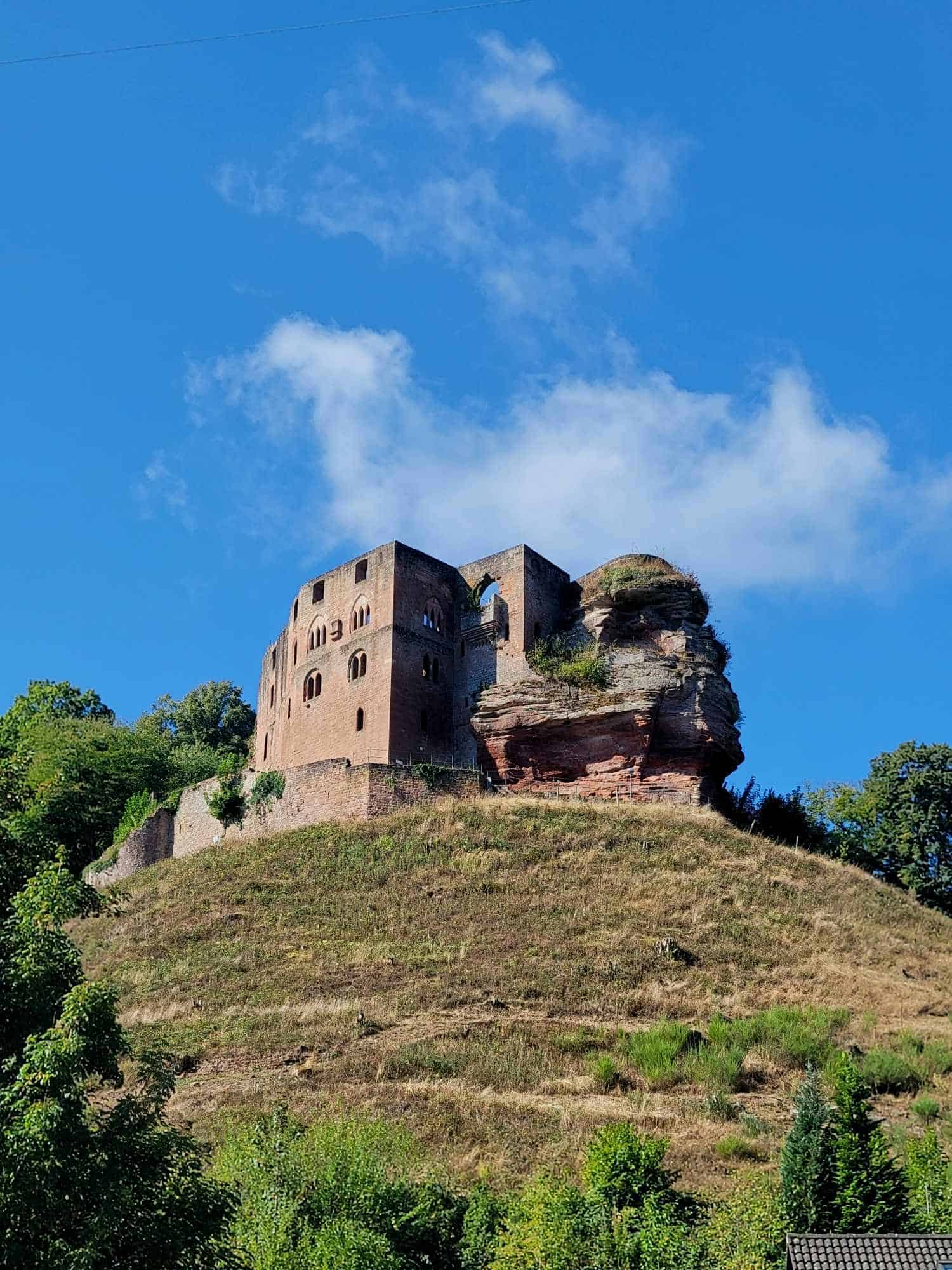 A large rectangular castle ruin made of red sandstone sits atop a grassy hill with blue sky and white clouds in the background