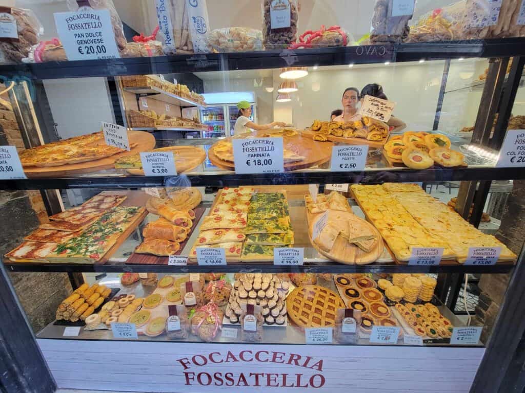 A storefront that reads Focacceria Fossatello and shelves up against the window with pastries and pies, small pizzask foccaccia bread, cookies and other baked goods