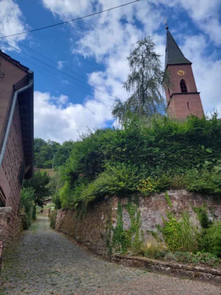 A stone wall covered with shrubbery next to a stone path leading up a hill. The spire of a church can be seen on the right