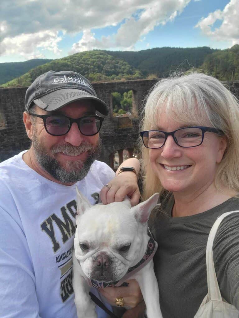 A smiling couple holding a french bulldog with castle ruin and forest in the background